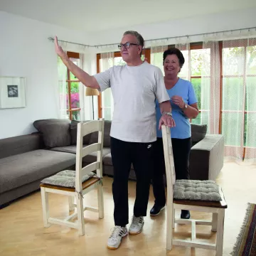 älterer Mann bei einer Bewegungsintervention in einer Wohnung mit einer älteren Dame